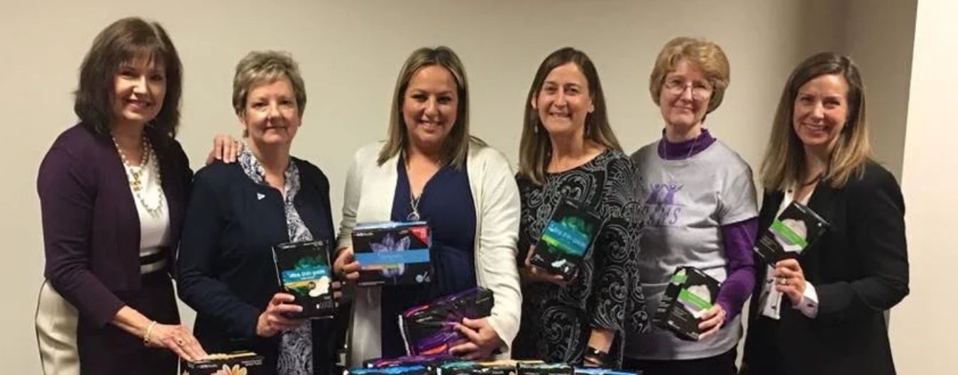 Six women stand behind a table displaying menstrual hygiene products, smiling during a community donation and awareness event.