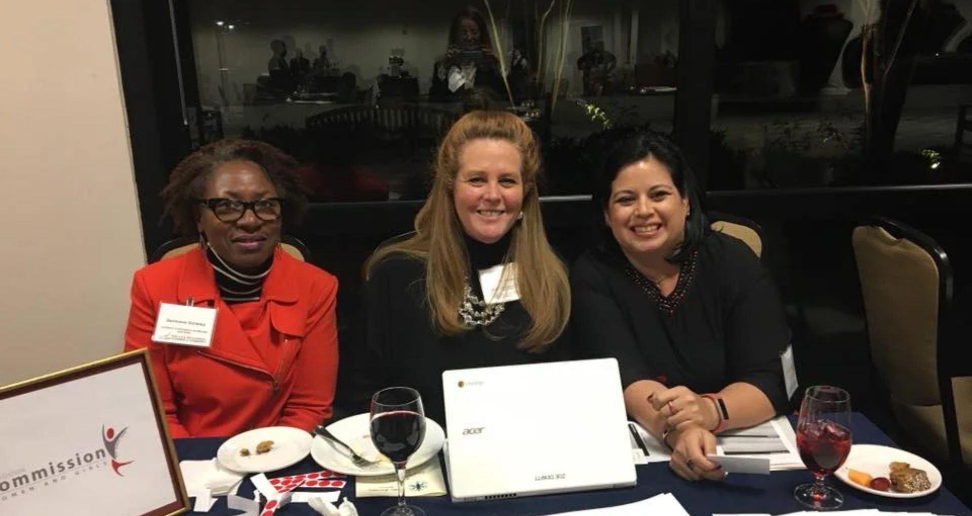 Three women seated at a table during a community event, smiling toward the camera with a laptop and refreshments in front of them.