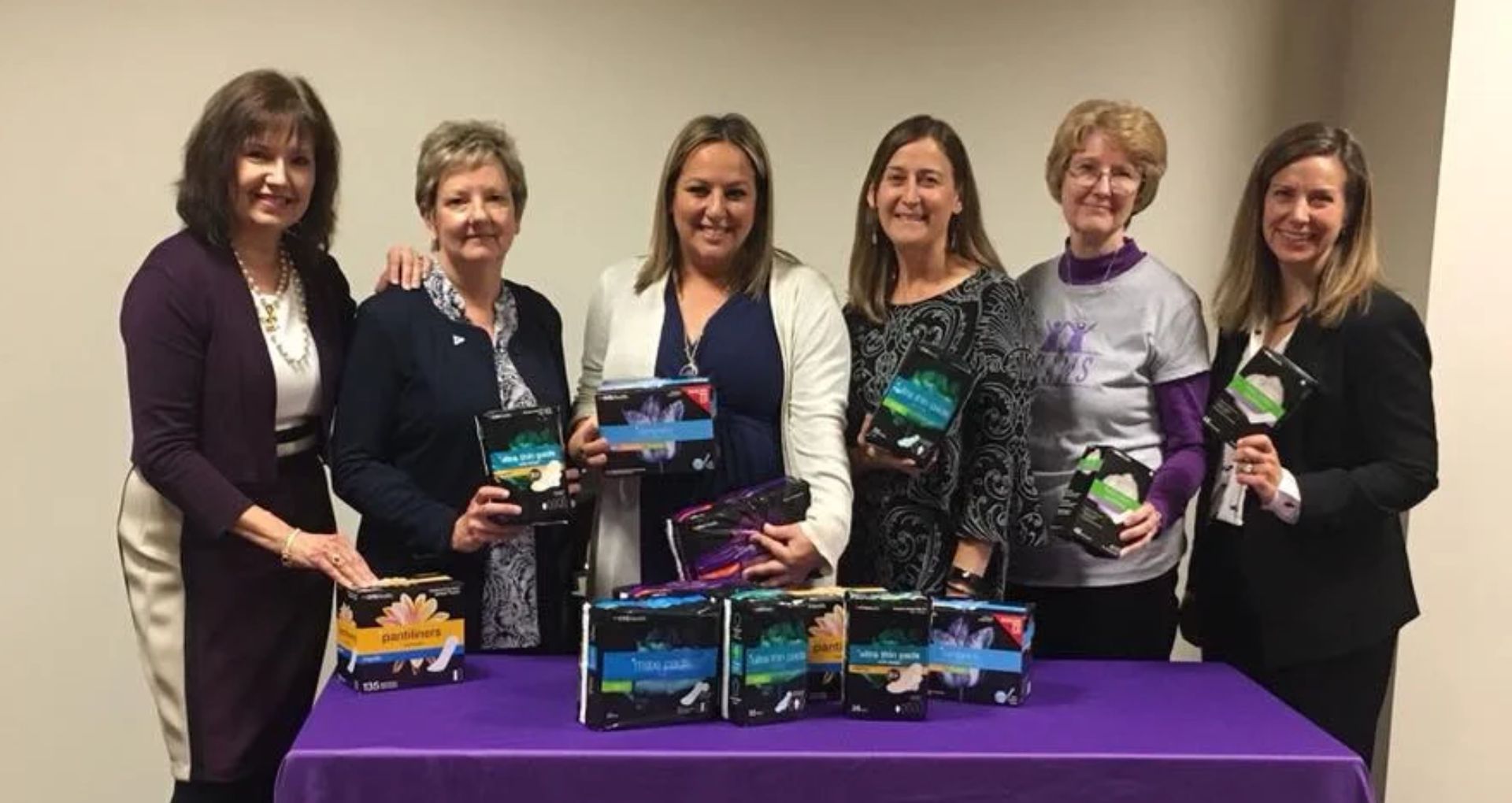 Six women stand behind a table displaying menstrual hygiene products, smiling during a community donation and awareness event.