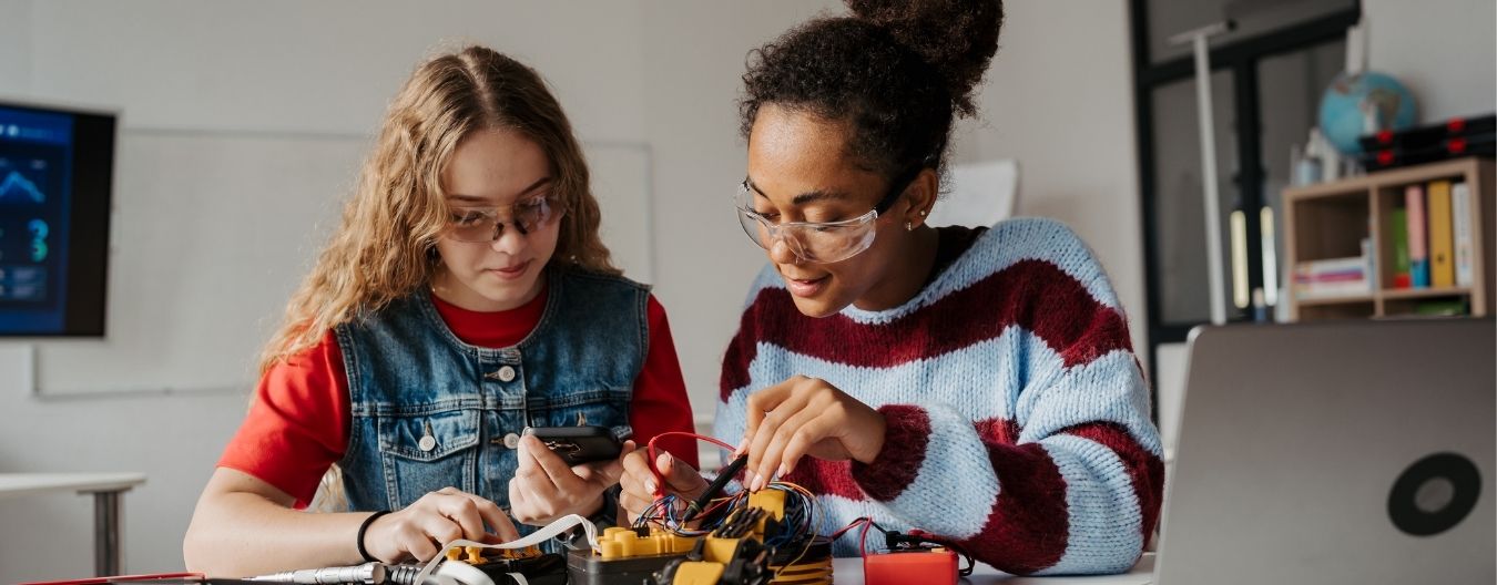Two students wearing safety goggles collaborate on a STEM project, working with electronics and wiring at a classroom table.