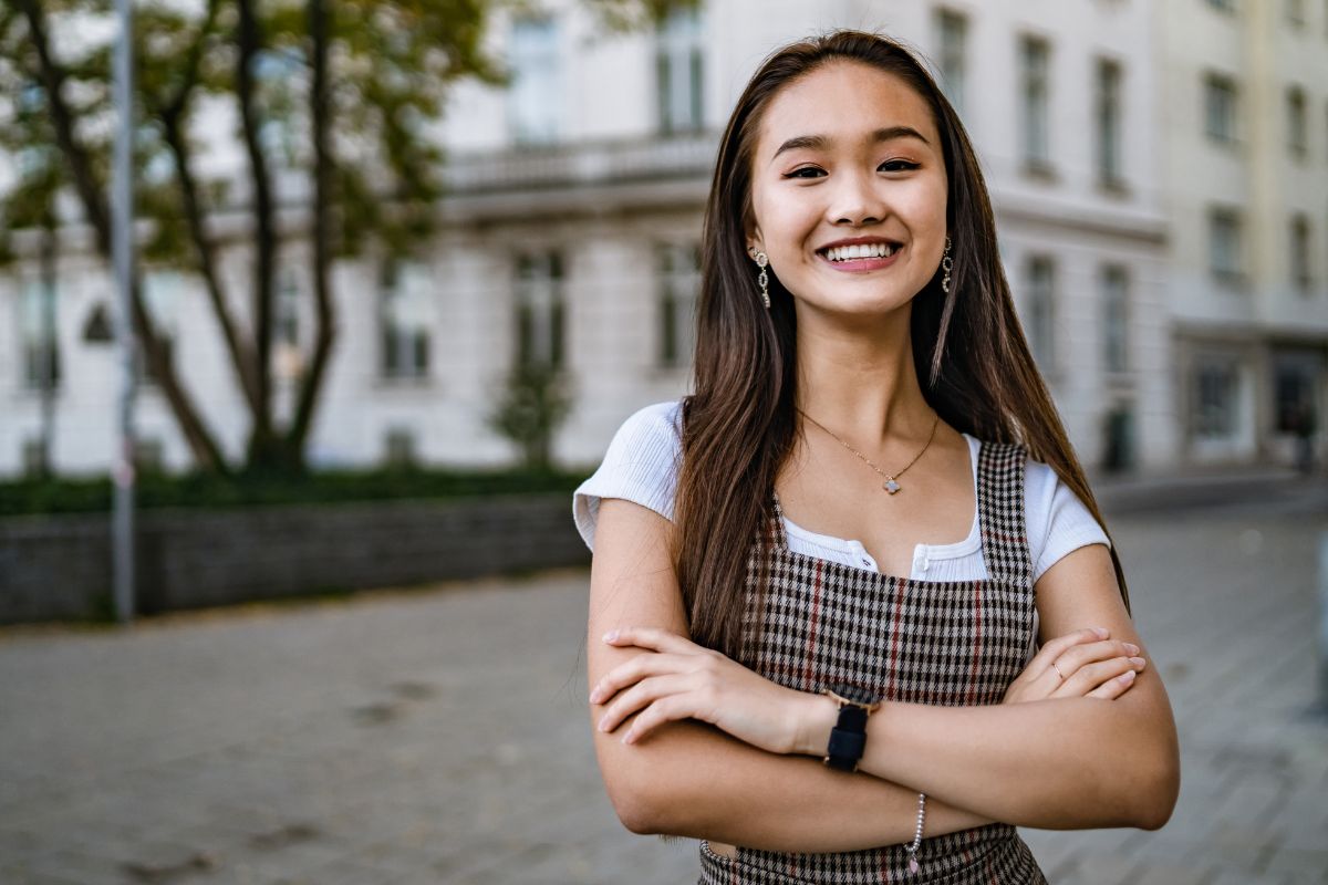 LCWAG Home Page Research & Impact Image LCWAG Home Page Research & Impact Image depicting a young Asian woman with her arms crossed smiling in a city setting.